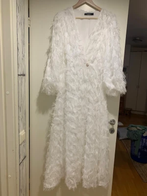 Vit fjädermönstrad lång kappa - Säljer en lång vit kappa från NA-KD med fjäderliknande struktur och en knapp framtill. Kappan har v-ringning och vida ärmar, vilket ger en cool och unik look. Perfekt för dig som vill sticka ut och gillar statement-plagg.