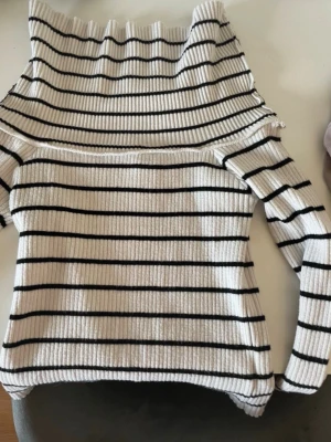 Randig stickad tröja från H&M - Säljer en vit ribbstickad tröja från H&M med svarta horisontella ränder och bred polokrage. Tröjan har lång ärm och är gjord i ett mjukt, stretchigt material som sitter snyggt på kroppen. Perfekt för en trendig och avslappnad look.