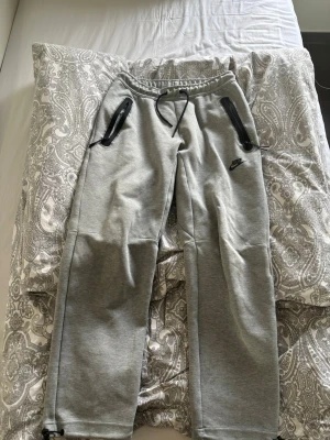 Gråa Nike mjukisbyxor med dragkedjor - Snygga grå mjukisbyxor från Nike med svarta dragkedjor på fickorna och justerbar midja med snörning. Perfekta för chill eller träning, med klassisk Nike-logga på låret och raka ben.
