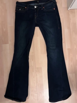 Mörkblå bootcut jeans True Religion - Mörkblå bootcut jeans från True Religion med klassiska kontrastsömmar och detaljerade bakfickor. Jeansen har låg midja och utsvängda ben. Lite slitna vid ena benet(se på bild)