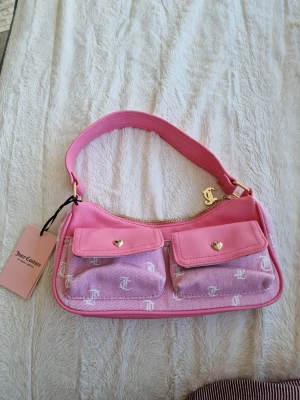 Rosa Juicy Couture handväska - Säljer en superfin rosa handväska från Juicy Couture med två fickor framtill och JC-logga i vitt över hela väskan. Väskan har guldfärgade detaljer, hjärtformade knappar och ett brett axelband. Materialet är en mix av tyg och konstläder. Perfekt för dig som gillar Y2K-stil!