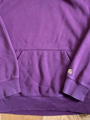 Lila hoodie från Carhartt - Snygg lila hoodie från Carhartt med klassisk känguruficka och ribbade muddar. Hoodien har en broderad Carhartt-logga på ärmen och är gjord i mjukt bomullsmaterial. Perfekt för en chill och avslappnad stil.