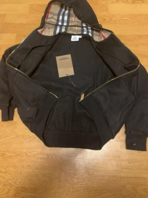 Svart hoodie med dragkedja från Burberry - Svart hoodie från Burberry med dragkedja och klassiskt rutigt foder i huvan. Hoodien har två fickor framtill, ribbade muddar och guldfärgad dragkedja. Perfekt för en stilren och lyxig streetlook.