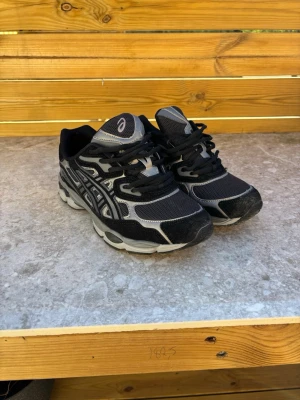Svarta Asics sneakers med grå detaljer - Snygga svarta sneakers från Asics med grå och silvriga detaljer. Skorna har klassisk mesh och mocka-look, samt tydliga Asics-loggor på sidorna. Sulan är vit och svart med sportig design. Perfekta för en streetwear-look eller till vardagsoutfiten. Använda max 3 gånger, 