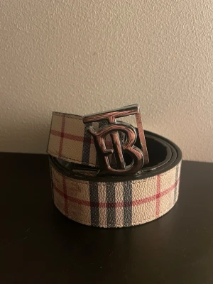 Burberry vändbart bälte med monogram - Stilrent vändbart bälte från Burberry med klassiskt rutigt mönster i beige, svart och rött på ena sidan och helsvart på andra. Snyggt metallspänne med Burberrys monogram. Perfekt accessoar för att lyfta din outfit.
