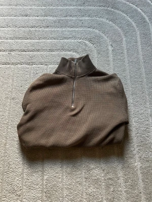 Brun stickad halvzip  - Snygg brun stickad tröja med halv dragkedja och hög krage. Tröjan har lång ärm, ribbade muddar och en avslappnad passform. Perfekt för lager-på-lager och enkel att matcha med jeans eller chinos.