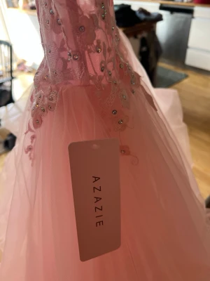 Rosa tyllklänning från Azazie - Säljer en ljusrosa klänning från Azazie med tyll och spetsdetaljer samt små glittrande stenar. Klänningen har en romantisk look med blommiga broderier upptill och flera lager tyg som ger volym.inte använd