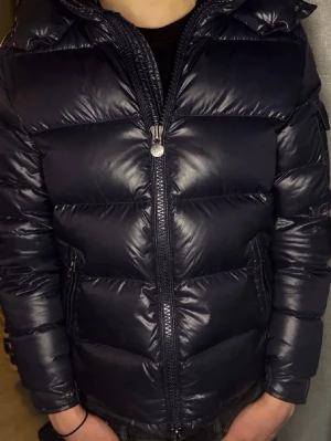 Mörkblå Moncler Maya - Snygg mörkblå dunjacka från Moncler med glansig finish, huva och dragkedja framtill. Jackan har breda kanaler, sidofickor med dragkedja och Moncler-logga på ärmen. Perfekt för kalla vinterdagar och riktigt trendig stil.