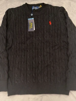 Svart kabelstickad tröja Polo Ralph Lauren - Kabelstickad svart tröja från Polo Ralph Lauren med rund halsringning och den klassiska röda logotypen broderad på bröstet. Tröjan har ribbade muddar vid ärmslut och nederkant. Perfekt för en stilren och tidlös look.