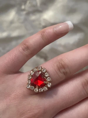 Ring med röd hjärtformad sten - Snygg ring med en stor röd hjärtformad sten i mitten, omgiven av glittrande klara stenar. Guldfärgad bas som ger en lyxig känsla. Perfekt för dig som vill sticka ut och addera lite glamour till din stil. Justerbar