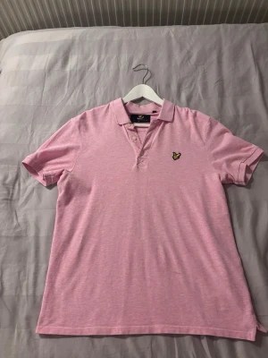 Rosa pikétröja från Lyle & Scott - Snygg rosa pikétröja från Lyle & Scott med klassisk krage, knäppning med tre knappar och den ikoniska gula loggan broderad på bröstet. Perfekt för en avslappnad men stilren look. Tillverkad i mjukt bomullsmaterial. Inga större täcken på användning! 