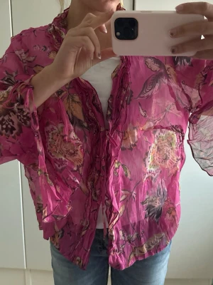 Rosa blommig transparent blus - Snygg rosa blus med blommigt mönster, volangkant längs knäppningen och snören vid ärmarna. Blusen är transparent och har långa, vida ärmar. Perfekt inför en sommardag med ett par jeans eller kjol!