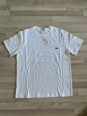 Lacoste T-shirt - Klassisk vit t-shirt från Lacoste med broderad krokodil-logga på bröstet. T-shirten har rund halsringning och korta ärmar. Tillverkad i mjuk bomull för en skön känsla och stilren look.