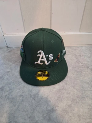 Grön New Era Oakland A's keps - Snygg grön keps från New Era med Oakland A's logga framtill. Kepsen har broderade fjärilar och detaljer på sidorna samt klassisk rak skärm. Perfekt accessoar för dig som gillar sportig och trendig stil.