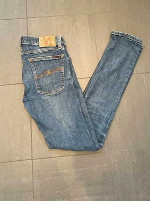 Blå jeans från Nudie Jeans, 28/32 - Snygga blå jeans från Nudie Jeans med klassisk femficksdesign och gul kontrastsöm. Jeansen har en rak passform och är tillverkade i denim av bomull. Bakfickan har Nudie Jeans karakteristiska broderi. Perfekta för en avslappnad stil.