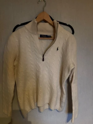 Vit kabelstickad tröja Polo Ralph Lauren - Kabelstickad vit tröja från Polo Ralph Lauren med halv dragkedja och hög krage. Tröjan har lång ärm och den klassiska broderade loggan på bröstet. Perfekt för lager-på-lager och en clean look. Står L men råka krympa den så den passar kanske är som är mellan 170-175 skull jag säga 