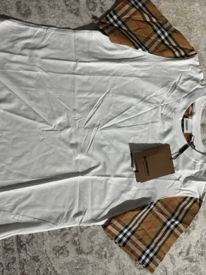 Vit Burberry t-shirt med rutiga ärmar - Snygg vit t-shirt från Burberry med klassiska beige, svarta och röda rutiga ärmar. T-shirten har rund halsringning och är tillverkad i mjukt bomullsmaterial. Perfekt för dig som gillar stilrena plagg med en twist.