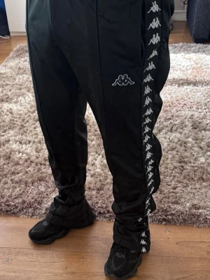 Svarta Kappa byxor med knappar XL - Säljer ett par svarta träningsbyxor från Kappa i storlek XL. Byxorna har vita Kappa-loggor längs båda benen och knappar längs sidorna. Resår i midjan för skön passform. Perfekta för sport eller chill.