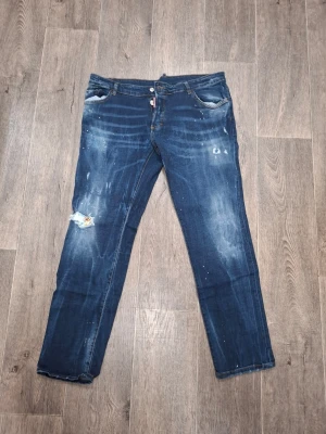 Blå distressed jeans från DSQ2 - Snygga blå jeans från DSQ2 med slitningar, färgstänk och hål på ena knät. Klassisk femficksmodell med läderpatch bak och cool tvätt. Tillverkade i Italien, perfekt för dig som gillar en edgy streetstil.
