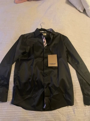 Svart skjorta från Burberry M - Snygg svart skjorta från Burberry i storlek M. Klassisk modell med långa ärmar och knappar framtill. Kragen har Burberrys ikoniska rutiga mönster på insidan för en extra detalj. Perfekt för dig som gillar stilrena plagg med exklusiv känsla. Använd endast en gång!