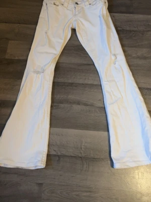 Vita bootcut jeans med slitningar - Säljer ett par vita bootcut jeans med slitningar vid knäna. Jeansen har klassisk femficksmodell och vida ben. Perfekta för dig som gillar en retrostil och vill sticka ut med ljusa jeans. Materialet känns som bomull med lite stretch. DOM ÄR UPPSYDDA MEN PASSAR MIG SOM ÄR 160 CM