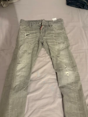 Grå distressed jeans från Dsquared2 - Säljer ett par grå jeans från Dsquared2 med slitna detaljer, broderade texter och färgstänk. Jeansen har rak passform, klassiska fem fickor och synlig logga vid gylfen. Perfekta för dig som gillar en edgy streetstil.