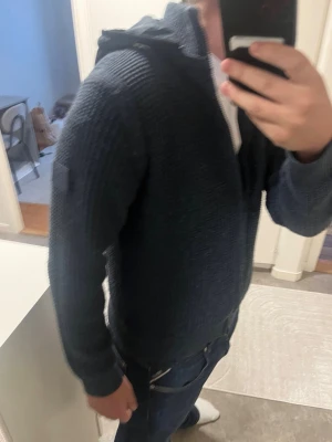 Mörkblå stickad hoodie med dragkedja - Säljer en mörkblå stickad hoodie med dragkedja framtill. Tröjan har huva, långa ärmar och en grovstickad struktur som ger en cool och avslappnad look. Perfekt för lager-på-lager och passar till jeans eller chinos.
