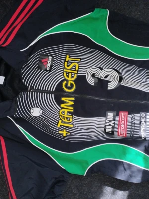 Adidas Teamgeist sportjacka svart/grön Ny m/L - Säljer en Adidas Teamgeist sportjacka i svart med gröna och vita detaljer samt röda ränder på axlarna. Jackan har dragkedja, tryckta loggor och text, samt nummer 3 på framsidan. Perfekt för fotbollsträning eller som streetwear.  Ny m/L rea 1000kr plus..