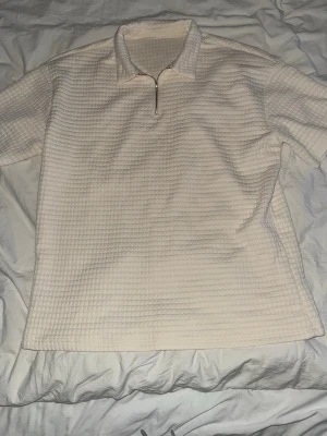 Vit ribbad polotopp med half zip - Vit polotopp med kort ärm och half zip-dragkedja framtill. Toppen har ett ribbat, rutigt mönster och är tillverkad i ett mjukt material. Perfekt för en stilren och avslappnad look.