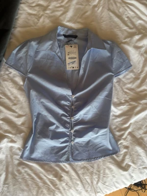 Ljusblå kortärmad blus från Zara - Snygg ljusblå blus från Zara med korta ärmar och klassisk krage. Blusen har knäppning framtill med små knappar och figurnära passform. Perfekt för en stilren och fräsch look. Materialet är tunt och lätt, vilket gör den bekväm att bära.