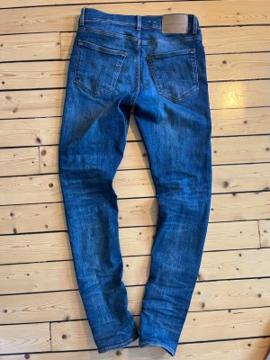 Blå slim fit jeans från Tiger of Sweden - Snygga blåa slim fit jeans från Tiger of Sweden med klassisk femficksmodell, storlek 28/32. Jeansen har en tvättad look med ljusa slitningar och har en skön passform. Ändå defekten är lädermärket på baksidan är lite slitet (se bild 4)