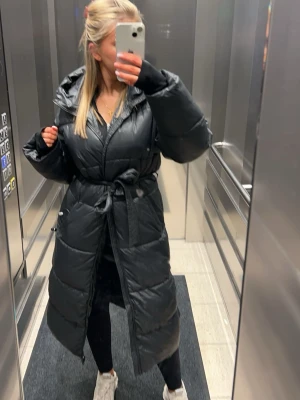 Svart lång pufferjacka med huva - Säljer en svart lång pufferjacka med huva och knytskärp i midjan. Jackan har en oversized passform, stora kanaler och är perfekt för kalla vinterdagar. Stängs med dragkedja och tryckknappar. Snygg och trendig modell som håller dig varm. Nypris 1000kr