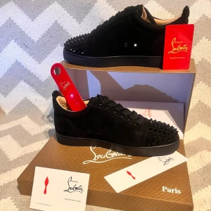 Svarta Louboutin sneakers med nitar - Snygga svarta sneakers från Christian Louboutin i mocka med svarta nitar på tån och sidorna. Skorna har klassisk röd sula och svarta skosnören. Låg modell som ger en clean och edgy look. Perfekta för dig som vill sticka ut med exklusiva detaljer. (Allt på bilderna medföljer)