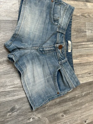 Blå denimshorts från Denimite - Klassiska blå jeansshorts från Denimite med fem fickor, bälteshällor och knappstängning. Shortsen har en ljus tvätt och normal passform, perfekta för varma dagar. Snygga detaljer med slitningar och synliga sömmar.