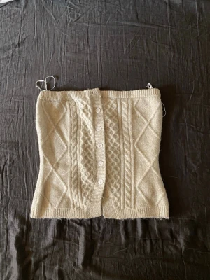 Beige stickad kortkjol med knappar - Trendig beige stickad kortkjol med kabelstickat mönster och dekorativa knappar framtill. Kjolen har ribbade kanter och ett klassiskt rutmönster i stickningen. Perfekt för att skapa en mysig och stilren look.