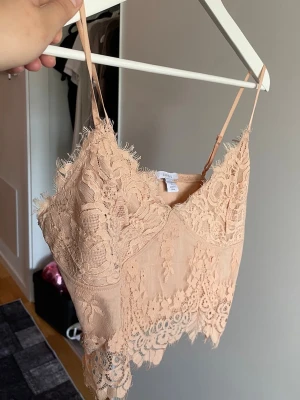 Beige spetslinnetopp från Amisu - Supersöt beige linnetopp från Amisu med tunna axelband och vacker spetsdetalj över hela toppen. Croppad modell med snygg v-ringning och dekorativt avslut nertill. Perfekt att styla med jeans eller kjol för en trendig look.