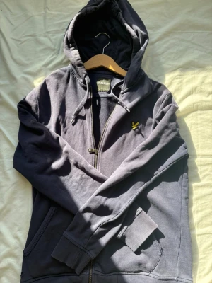 Mörkblå hoodie från Lyle & Scott - Har litet hål bild 3. Snygg mörkgrå hoodie från Lyle & Scott med dragkedja, huva med dragsko och klassisk gul logga på bröstet. Mjukt bomullsmaterial och två fickor framtill. Perfekt för en chill och avslappnad stil.