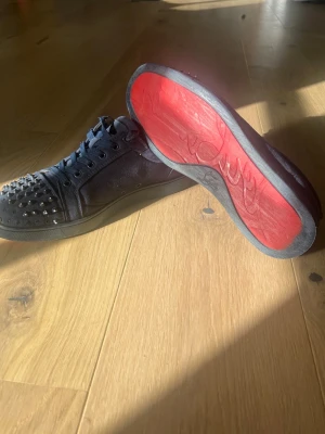 Christian lovotin - Säljer ett par svarta sneakers från Christian Lovotin med ikoniska röda sulor och coola nitar på tån. Skorna har lågt skaft, snörning och är tillverkade i mocka och läder. Perfekta för dig som vill sticka ut med en edgy och lyxig stil.Vissa spikar är lite lösa och sulan har gått upp lite.Skon är alltså inte äk….Skriv om ni är intresserade.
