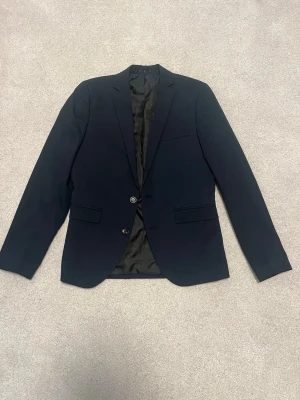 Svart kavaj från Jack & Jones - Stilren svart kavaj från Jack & Jones med två knappar framtill och klassiska fickor med lock. Kavajen har slät finish och helsvart foder, perfekt för en dressad look. Materialet är mjukt och följsamt för en snygg passform. Använd en kväll bara