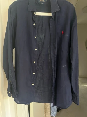 Mörkblå linneskjorta från Polo Ralph Lauren - Snygg mörkblå skjorta från Polo Ralph Lauren i linne med klassisk krage och vita knappar. Skjortan har lång ärm och den ikoniska röda logotypen broderad på bröstet. Perfekt för en stilren och avslappnad look.
