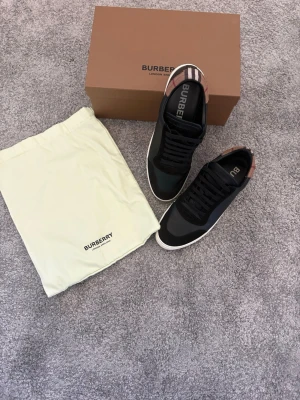 Svarta Burberry sneakers med rutdetalj - Snygga svarta sneakers från Burberry med klassiska beige och röda rutdetaljer på hälen. Skorna har en låg siluett, svarta skosnören och vit sula. Tillverkade i en mix av läder och textil för en exklusiv känsla. Perfekta för dig som gillar stilrena och trendiga skor. Box ingår, dustbag och kvitto finns. Pris kan alltid diskuteras!!!