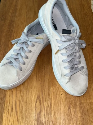 Beige Adidas Sleek sneakers i mocka - Snygga beiga Adidas Sleek sneakers i mocka med klassisk låg profil. Skorna har vita sulor, ljusgrå skosnören med gulddetaljer och diskret Adidas-logga på plösen. Perfekta för en clean och stilren look.