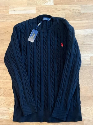 Svart kabelstickad tröja Polo Ralph Lauren - Svart kabelstickad tröja från Polo Ralph Lauren med rund halsringning och röd broderad logga på bröstet. Tröjan har långa ärmar och ribbade muddar vid ärmslut och nederkant. Klassisk och tidlös design i mjukt material.