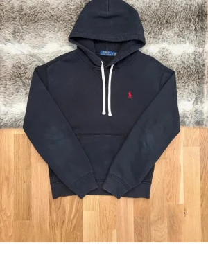 Svart hoodie från Polo Ralph Lauren - Klassisk svart hoodie från Polo Ralph Lauren med vit dragsko och röd broderad logga på bröstet. Hoodien har känguruficka framtill och ribbade muddar vid ärmar och nederkant. Perfekt för en avslappnad och stilren look. Snören fins tyvärr inte 