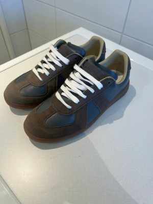 Bruna och blå gats med mockadetaljer - Snygga gats i brunt och mörkblått med ovandel i mocka och läder. Skorna har vita snören, beige insida och en klassisk gummisula. Perfekt för dig som gillar stilrena och tidlösa sneakers med retrovibbar. Skorna är endast använda en gång och säljer pågrund av att dom är för små