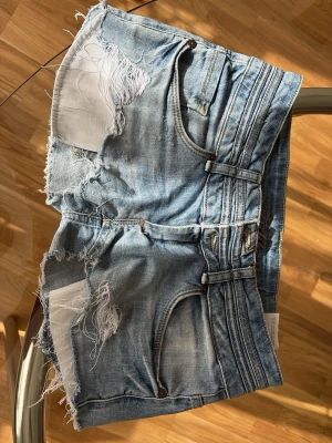 Blåa jeansshorts från Fishbone - Midja 39 Rise 17 Storlek 27 Snygga blå jeansshorts från Fishbone med råa kanter och slitna detaljer. Shortsen har hög midja, klassiska fem fickor och dubbla knappar framtill. Perfekta för en avslappnad och trendig look i sommar.
