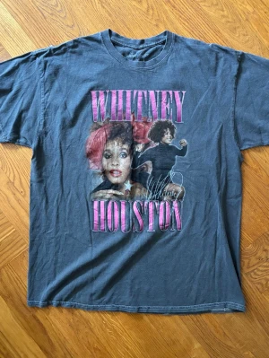 Whitney Houston tour t-shirt 2010 vintage  - Säljer en grå vintage t-shirt med stort Whitney Houston-tryck fram och turnédatum på ryggen. T-shirten har korta ärmar, rund hals och är gjord i mjuk bomull. Perfekt för dig som älskar vintage band merch och vill sticka ut med en ikonisk look.