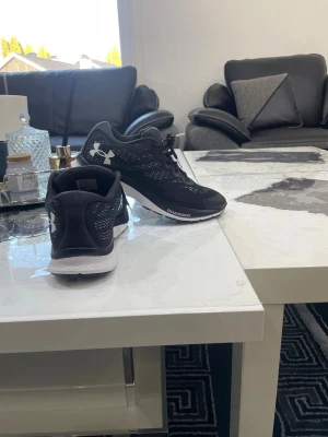 Svarta Under Armour sneakers - Snygga svarta sneakers från Under Armour med vit sula och klassisk logga på sidan. Skorna har meshdetaljer för extra andningsförmåga och snörning framtill. Perfekta för en sportig och stilren look.