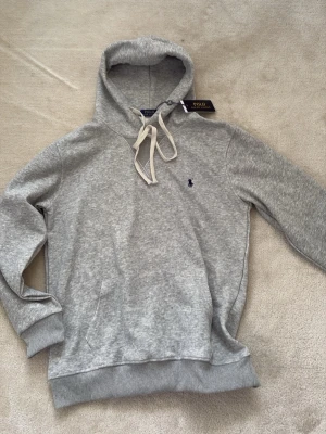 Grå hoodie från Polo Ralph Lauren - Snygg grå hoodie från Polo Ralph Lauren med klassisk huva och vita dragsnören. Tröjan har ribbade muddar, magficka och den ikoniska lilla loggan broderad på bröstet. Tillverkad i mjukt bomullsmaterial som känns riktigt skönt mot huden.