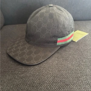 Gucci keps - Snygg keps från Gucci i mörkgrått med klassiskt GG-monogram över hela ytan. Kepsen har en böjd skärm och en ikonisk grön och röd rand på sidan. Tillverkad i canvasmaterial och har märkets logga på insidan. Perfekt för dig som gillar exklusiva accessoarer.